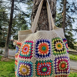 May include: A large, crocheted tote bag with a cream background and colorful granny square designs. The bag has two long straps and is hanging from a tree trunk.