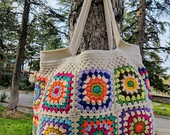 Op de afbeelding: Een grote, gehaakte tote bag met een crèmekleurige achtergrond en kleurrijke granny square-ontwerpen. De tas heeft twee lange hengsels en hangt aan een boomstam.