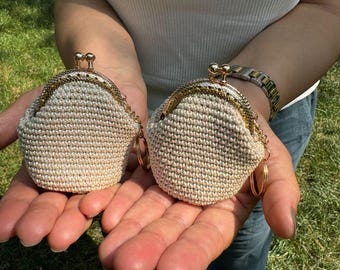 May include: Two small crocheted coin purses with gold clasps and chain accents. The purses are beige and have a textured woven design.