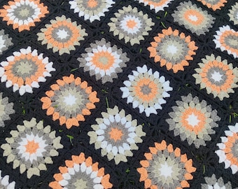 May include: A handmade crochet blanket featuring a repeating floral pattern. The blanket's design incorporates black, white, gray, and orange squares. The floral motifs are centered with white and surrounded by gray, orange, and black. The blanket is likely made of soft yarn.