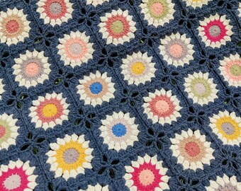 May include: A crocheted blanket with a blue background and colorful floral patterns. The blanket is made of yarn and has a textured surface.