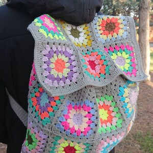 Op de afbeelding: Een grijze gehaakte rugzak met kleurrijke granny squares. De rugzak heeft een verscheidenheid aan kleuren, waaronder roze, groen, geel en blauw.