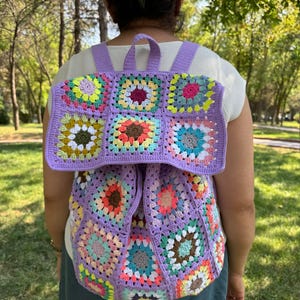 May include: A purple crochet backpack with a colorful granny square pattern. The backpack has adjustable straps and a zipper closure.