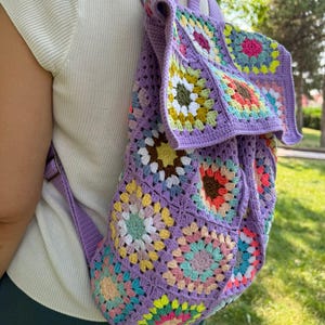 May include: A purple crochet backpack with a colorful granny square pattern. The backpack has a drawstring closure and adjustable straps.