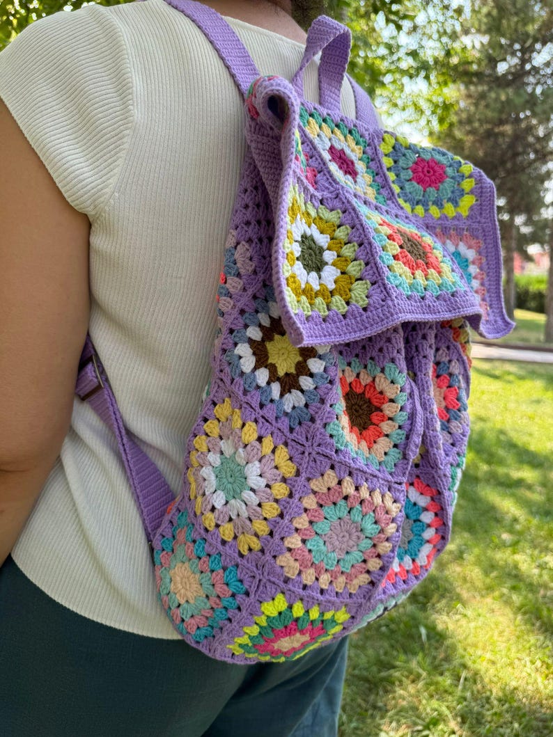 May include: A purple crochet backpack with a colorful granny square pattern. The backpack has a drawstring closure and adjustable straps.