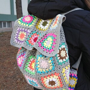 Op de afbeelding: Een grijze gehaakte rugzak met kleurrijke granny squares. De rugzak heeft een trekkoord sluiting en verstelbare banden.