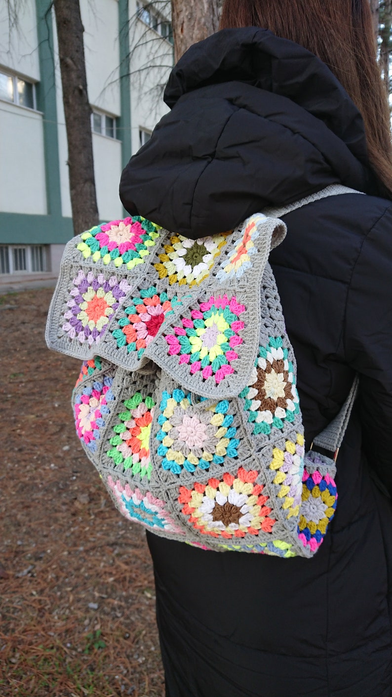 Op de afbeelding: Een grijze gehaakte rugzak met kleurrijke granny squares. De rugzak heeft een trekkoord sluiting en verstelbare banden.
