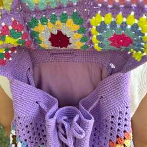 May include: A purple crochet backpack with a colorful granny square pattern. The backpack has a drawstring closure and is made of soft yarn.