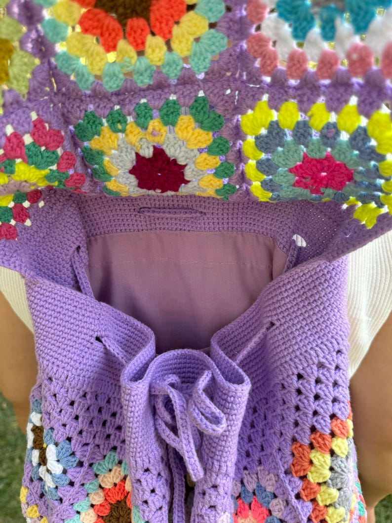 May include: A purple crochet backpack with a colorful granny square pattern. The backpack has a drawstring closure and is made of soft yarn.