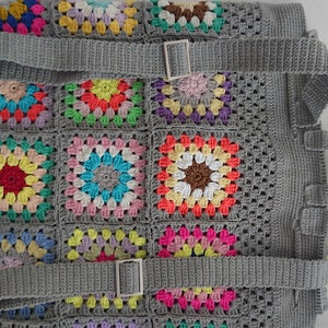 Op de afbeelding: Een grijze gehaakte rugzak met kleurrijke granny square-patches en verstelbare banden. De banden hebben zilveren gespen.