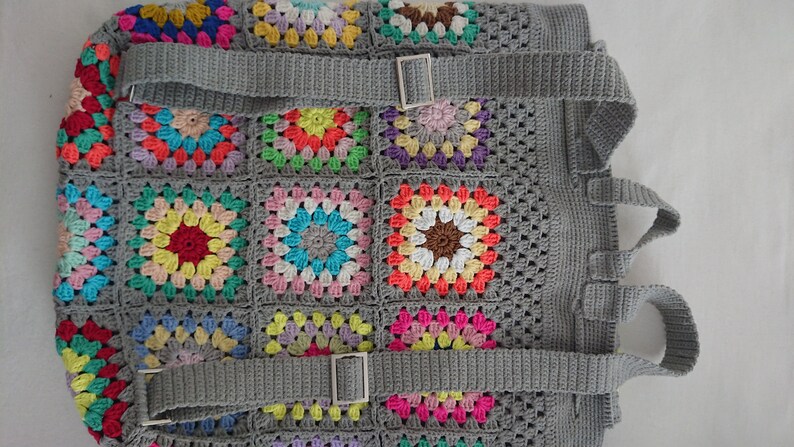 Op de afbeelding: Een grijze gehaakte rugzak met kleurrijke granny square-patches en verstelbare banden. De banden hebben zilveren gespen.