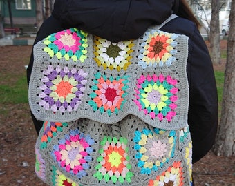 Può includere: Uno zaino in crochet grigio con quadrati di granny colorati. Lo zaino è realizzato con una varietà di colori vivaci, tra cui rosa, giallo, verde, blu e arancione.