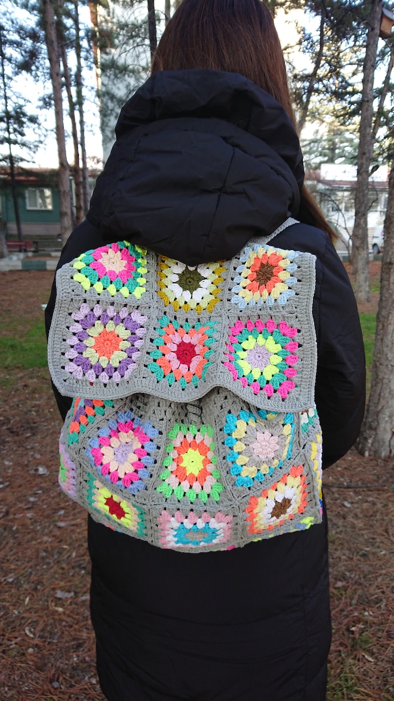 Op de afbeelding: Een grijze gehaakte rugzak met kleurrijke granny squares. De rugzak is gemaakt met een verscheidenheid aan felle kleuren, waaronder roze, geel, groen, blauw en oranje.