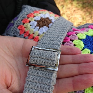 Op de afbeelding: Een grijze gehaakte tas met een zilveren gesp. De tas is gemaakt met kleurrijke granny squares in tinten roze, paars, groen en blauw.