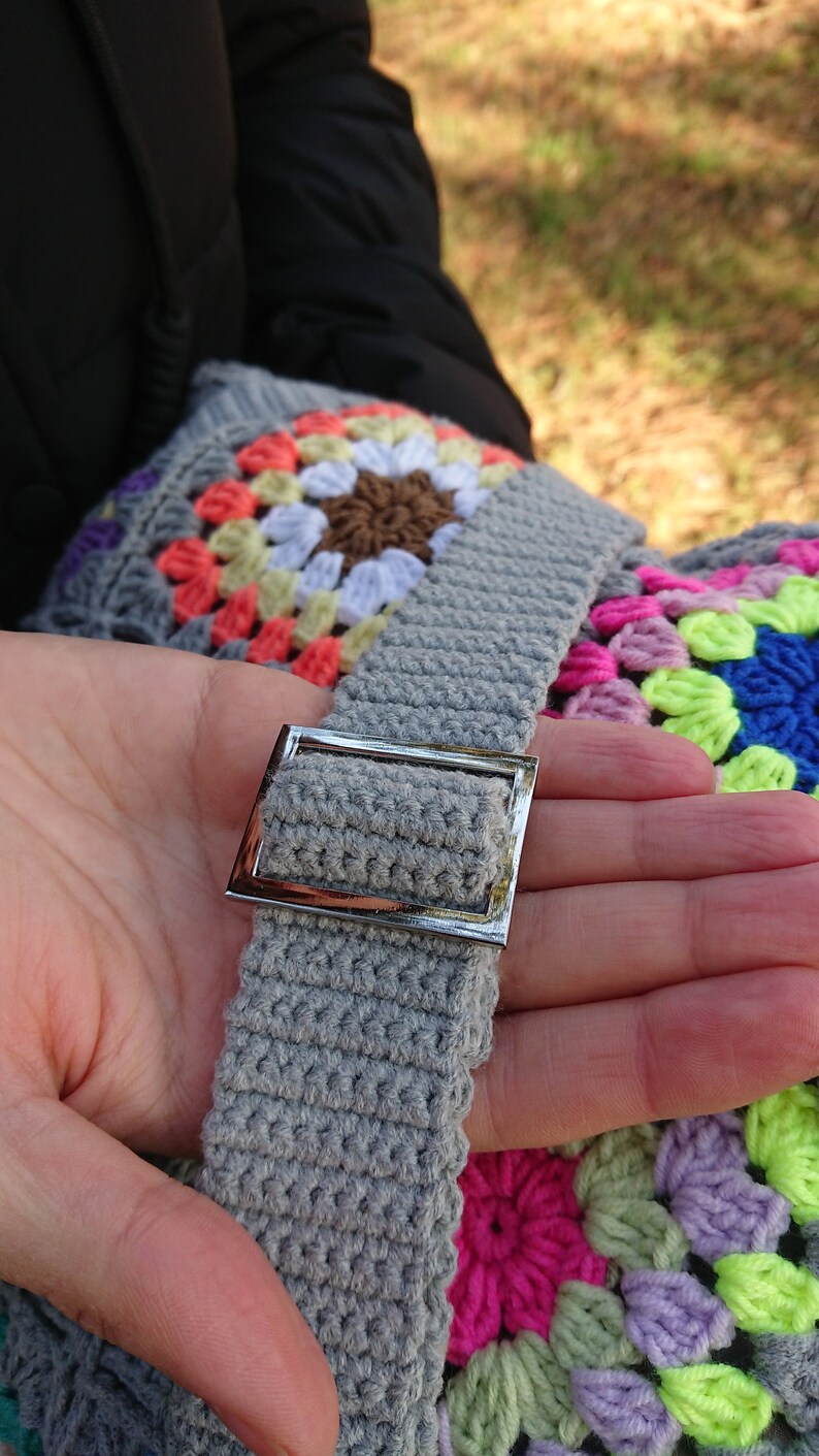 Op de afbeelding: Een grijze gehaakte tas met een zilveren gesp. De tas is gemaakt met kleurrijke granny squares in tinten roze, paars, groen en blauw.