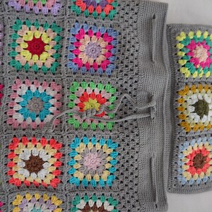 Op de afbeelding: Een grijze gehaakte tote bag met een trekkoord. De tas is gemaakt van granny squares in verschillende kleuren, waaronder roze, blauw, groen, geel en rood.
