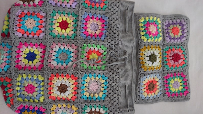 Op de afbeelding: Een grijze gehaakte tote bag met een trekkoord. De tas is gemaakt van granny squares in verschillende kleuren, waaronder roze, blauw, groen, geel en rood.