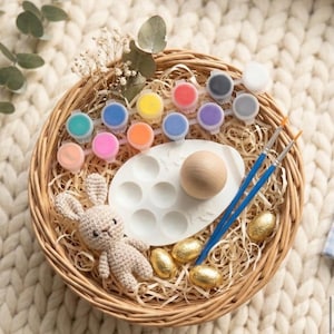 May include: A round wicker basket containing craft supplies. It features small paint pots in various colours, a wooden egg, a white paint palette, two blue paintbrushes, a crocheted bunny, and gold-wrapped chocolate eggs. The basket rests on a cream knitted blanket.