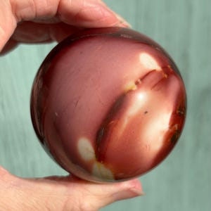 May include: A polished, spherical stone with a mix of colours. The stone features a base of deep red and burgundy, with streaks of white and cream. The surface is smooth and reflective, with a natural, organic appearance.