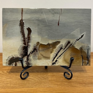 May include: A rectangular slab of polished picture jasper stone featuring a landscape-like pattern of grey, beige, and cream tones with dark brown, dendritic fern-like inclusions. The stone is displayed on a black metal scrollwork stand.