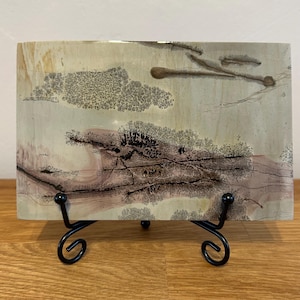 May include: A rectangular slab of polished dendritic jasper stone featuring intricate, tree-like black and brown mineral inclusions against a smooth, pale beige and soft pink background, displayed on a black metal scrollwork display stand.