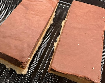 May include: Two rectangular caramel shortbread bars featuring a thick layer of smooth milk chocolate on top, a golden caramel center, and a crumbly shortbread base, arranged side by side on a black ridged plastic display tray with crumbs.