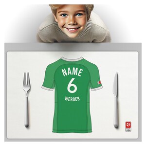 Puede incluir: Un mantel individual verde con el tema de una camiseta de fútbol con el nombre y el número 6. La palabra "WERDER" está debajo del número. Un tenedor y un cuchillo están a cada lado del mantel individual. Un niño está en el fondo.