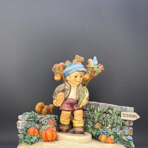 May include: A ceramic figurine depicting a young boy in autumn attire, holding a book and standing near a stone wall with a "School" sign. The scene includes pumpkins, a squirrel, and a tree with apples. The base reads "Autumn".