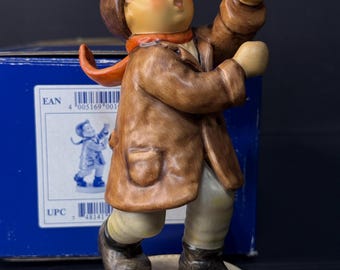 Może przedstawiać: Ceramiczna figurka śpiewającego dziecka, ubranego w brązowy płaszcz, czapkę i buty, z czerwoną szarfą. Figurka stoi na okrągłej, kremowej podstawie. Ręce dziecka są uniesione, a usta otwarte.