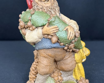 Könnte beinhalten: Eine texturierte Figur eines lächelnden Gnoms mit roter Mütze, blauer Weste und brauner Hose. Der Gnom hält ein Bündel aus grünen Blättern und braunen Eicheln. Der Sockel zeigt den geprägten Text CHEER und Clark 1996, umgeben von Eicheln.