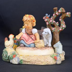 May include: A ceramic figurine featuring a child with a floral headband, wearing a brown vest and pink skirt, kneeling on a base. A white chicken, a small white lamb, a flowering tree with a blue bird, and a watering can are included.
