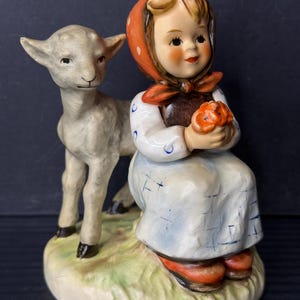 May include: A ceramic figurine depicting a young girl in a white dress and red headscarf, holding a flower, with a lamb standing beside her. The figurine has a painted finish with details like the girl's rosy cheeks and the lamb's fur.