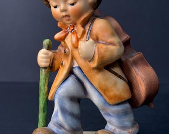 May include: A ceramic figurine of a traveler wearing a dark gray hat, a tan jacket with an orange neckerchief, and blue trousers. The figure carries a brown stringed instrument on the back and holds a green walking stick while standing on a base.