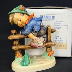 May include: A ceramic figurine of a child in a green hat and blue jacket climbing a brown wooden fence, with a small green frog at the base. The box displays the text In tausend Ängsten, Retreat to Safety, # 832, Hum 201/2/0, and 10 cm/4 inch.