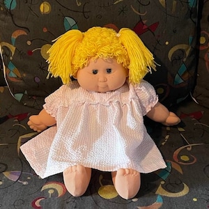 Puede incluir: Una muñeca vintage con coletas de hilo amarillo y un vestido rosa de seersucker. La muñeca tiene una cara redonda con pecas y lleva un vestido rosa claro con un patrón de seersucker blanco. Los brazos y las piernas de la muñeca son de un material suave.