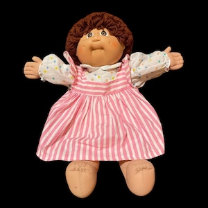 Puede incluir: Una muñeca vintage con una cara redonda, cabello de hilo marrón y ojos grandes. La muñeca viste un vestido a rayas rosas y blancas con cuello blanco y mangas salpicadas de círculos pastel. Los brazos de la muñeca están extendidos.