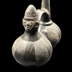 Puede incluir: Dos vasijas de cerámica gris oscuro con cuerpos redondeados y cuellos largos. Una vasija presenta una cara esculpida con un sombrero. Ambas vasijas tienen patrones geométricos tallados en sus superficies, sobre un fondo negro.