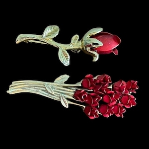 May include: Two gold-toned rose brooches with red enamel detailing. One brooch features a single rose bud on a stem with leaves. The other is a bouquet of red roses, also with gold-toned stems and leaves. Both are set against a black background.