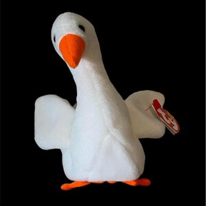 May include: A white plush goose toy with an orange beak and feet. The goose has outstretched wings and a red tag attached. The toy is set against a black background.