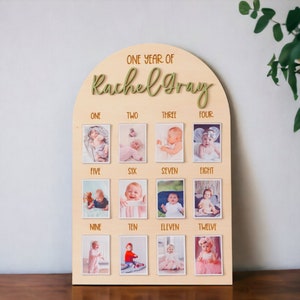 May include: A wooden photo display with a light brown background and a light brown arch at the top. The arch has the text "ONE YEAR OF Rachel Gray" in green lettering. The display has 12 photo slots, each with a number from one to twelve. Each slot has a photo of a baby girl.