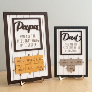 Könnte beinhalten: Zwei gerahmte Holzschilder mit den Worten "Papa" und "Dad" in Schreibschrift. Jedes Schild hat einen weißen Hintergrund mit schwarzen und braunen Rahmen. Die Schilder enthalten den Text "YOU ARE THE PIECE THAT HOLDS US TOGETHER" und personalisierte Puzzleteile.