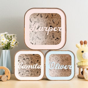 Puede incluir: Tres cajas de sombra cuadradas de madera con marcos de colores claros en rosa, blanco y azul. Cada caja cuenta con un panel frontal transparente con un nombre personalizado en letra blanca: Harper, Camila y Oliver. Los fondos muestran patrones de flores, dinosaurios y temas espaciales.