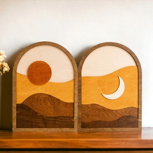 Pu&ograve; includere: Due opere d'arte da parete in legno raffiguranti una scena di sole e luna. Il pezzo del sole presenta un sole rotondo arancione e un paesaggio a strati nei toni del marrone e del giallo. Il pezzo della luna mostra una luna crescente e un paesaggio simile.