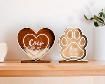 May include: Two wooden pet memorial keepsakes. One is a heart-shaped box with the name "Coco" and the dates "2018 - 2024" engraved on it. The other is a paw print-shaped box with the name "Ace" and the dates "2020 - 2021" engraved on it.