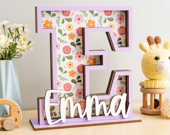 Puede incluir: Una letra E de madera púrpura con un patrón floral en su interior. La letra está de pie sobre una base con el nombre "Emma" escrito en blanco.