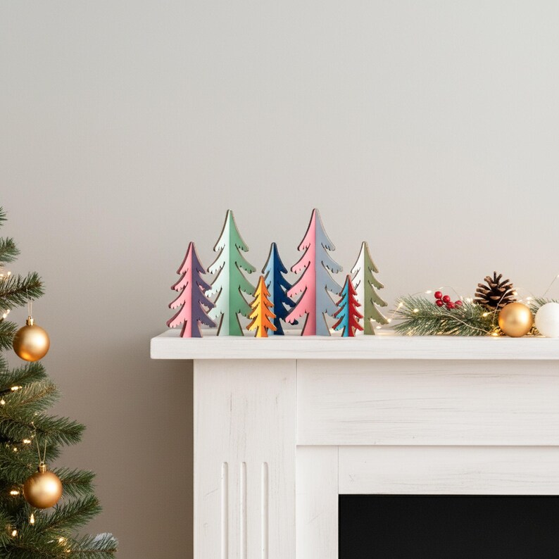 May include: A collection of seven small, flat, wooden Christmas tree ornaments in various colours including purple, light green, orange, dark blue, pink, red, and white, arranged in a row on a white mantelpiece with a pine garland.