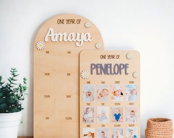 Puede incluir: Dos tableros de hitos para bebés de madera. Un tablero arqueado con el texto "ONE YEAR OF Amaya" y detalles de margaritas. El otro tablero tiene el texto "ONE YEAR OF Penelope" y ranuras para fotos para cada mes.