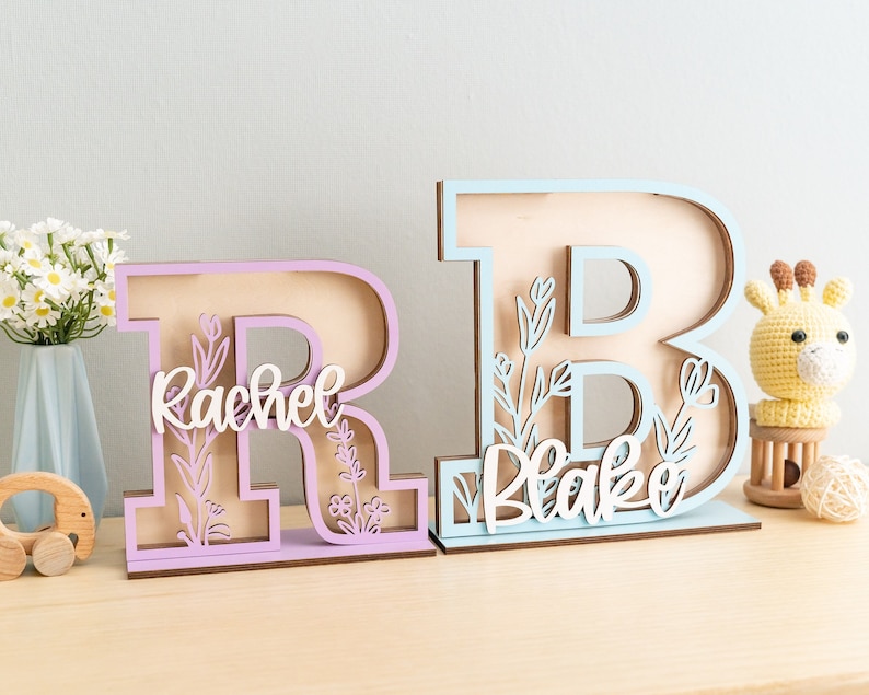 May include: Two wooden letters, one light blue and one light purple, stand on a wooden surface. The letters are decorated with floral cutouts and the names "Blake" and "Rachel" are cut out of the wood.