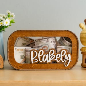 May include: A wooden money box with a clear window, filled with US dollar bills. The box has the name "Blakely" written on it in white letters.