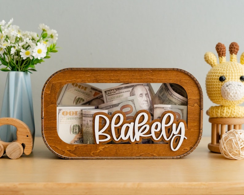 May include: A wooden money box with a clear window, filled with US dollar bills. The box has the name "Blakely" written on it in white letters.